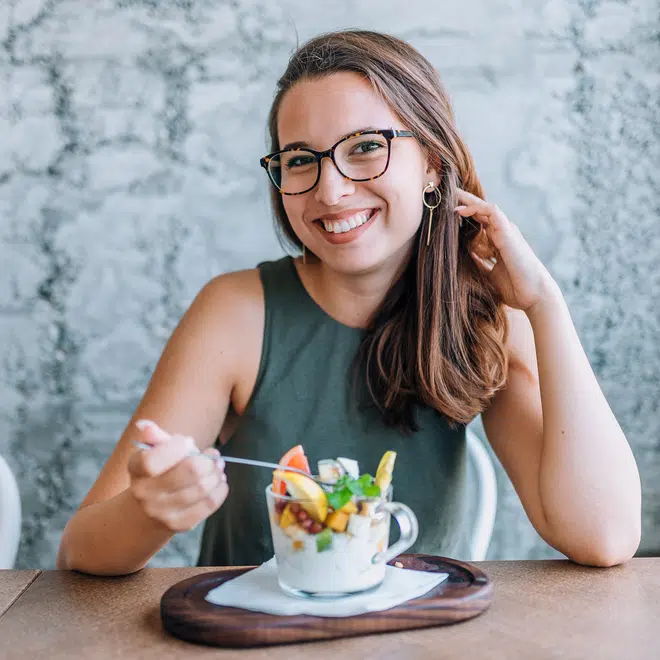 smiling woman in glasses sitting down with a spoon in her hand and a yogurt parfait with fresh fruit on a table in front of her