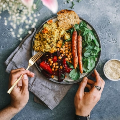 hands holding a fork near a bowl of food with greens, carrots, legumes, and bread