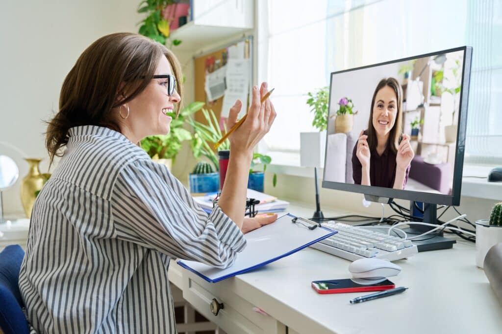 A woman sitting at a desk with a clipboard in front of her deskktop computer and virtually talking to another woman