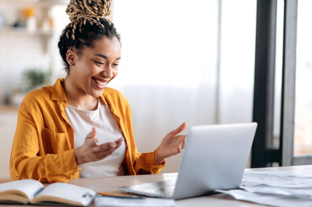 A woman sitting at a table and lifting her hands while smiling at her open laptop