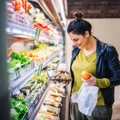 woman in the produce aisle of a grocery store holding an apple and package of mushrooms