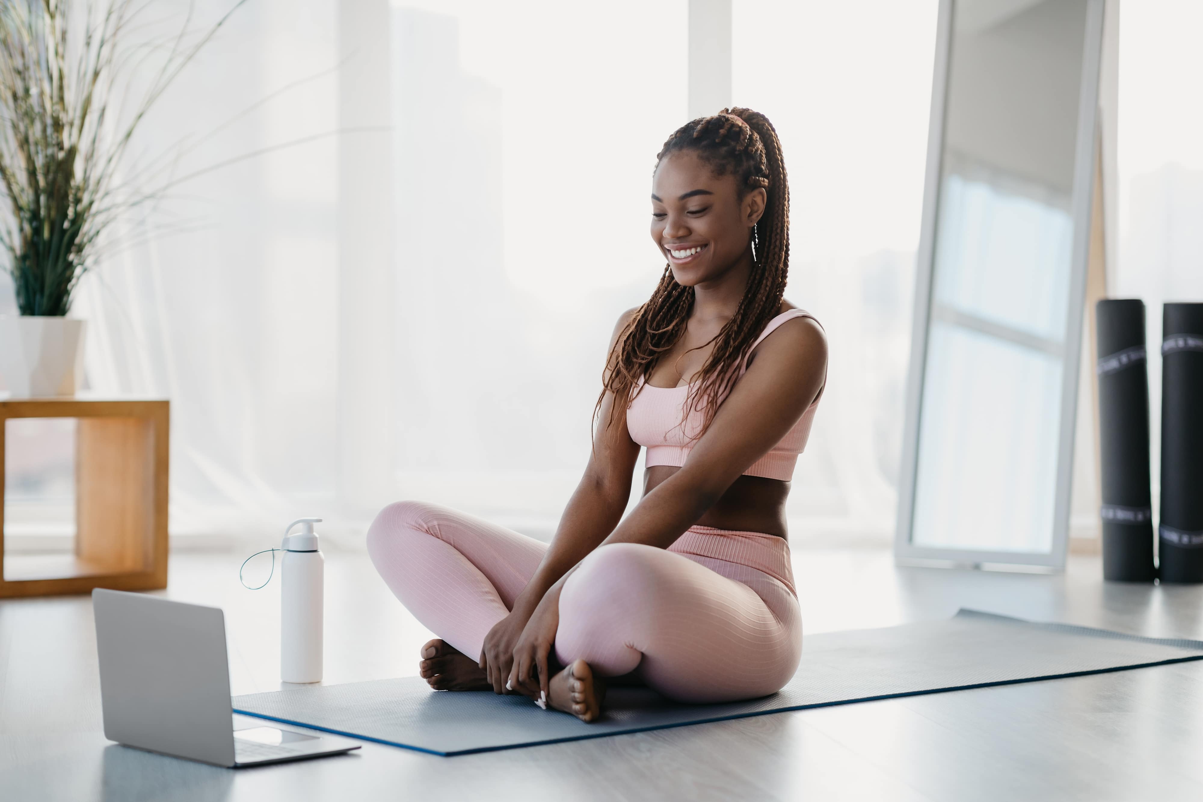 woman in pink athletic wear sitting on a yoga mat in front of her laptop and smiling